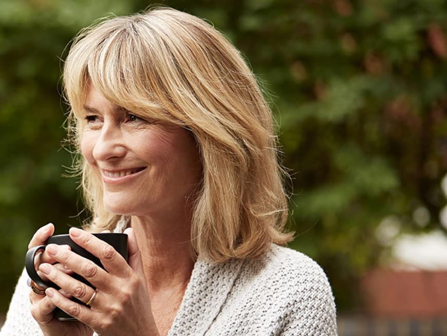 Blonde Frau mittleren Alters mit Kaffeetasse in der Hand steht lächelnd im Garten