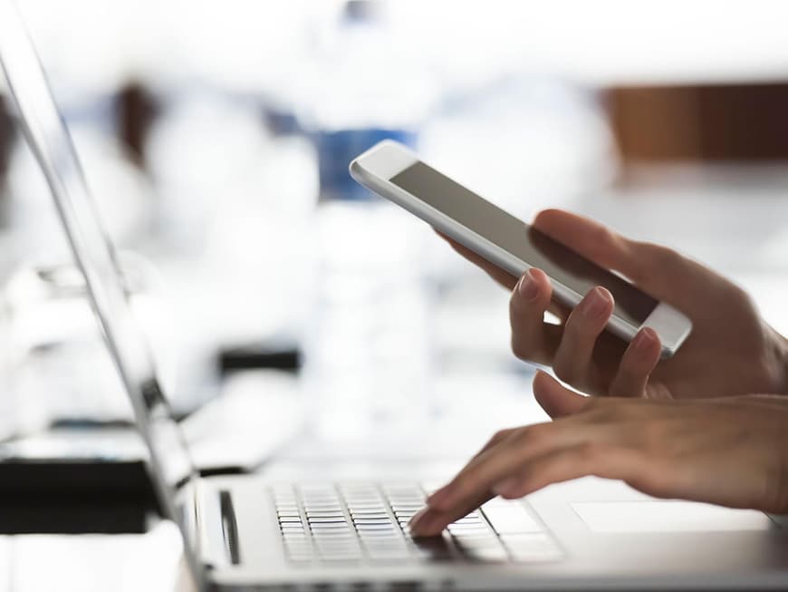 Hand auf der Tastatur eines Laptops, in der anderen Hand ein Smartphone