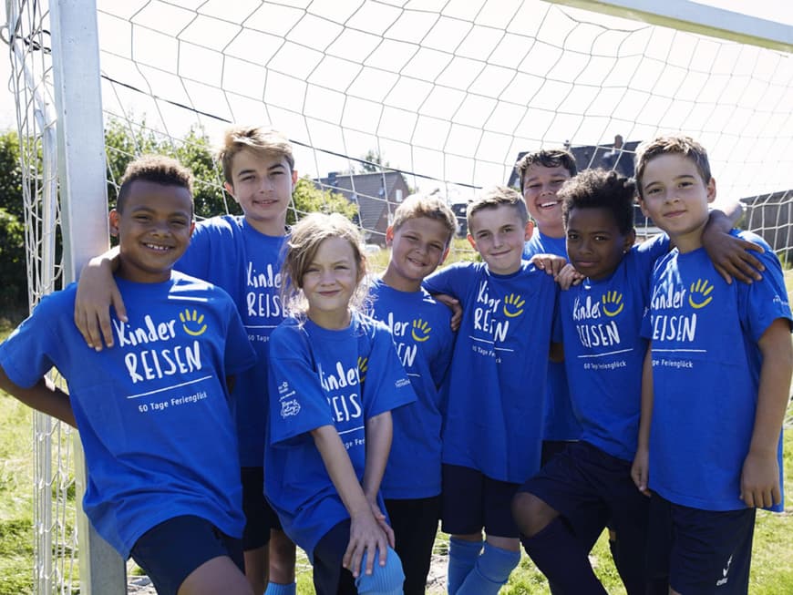 Acht Kinder in blauen T-Shirts mit der Aufschrift Kinderreisen vor einem Fußballtor