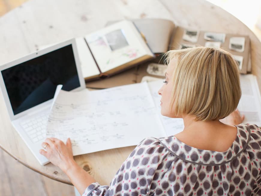 Blonde Frau sitzt am Tisch vor Laptop und schriftlichen Aufzeichnungen vom Stammbaum sowie Fotoalben