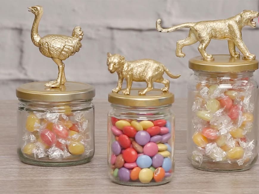 Drei Einmachgläser mit goldenen Deckeln und Tierfiguren darauf, befüllt mit Bonbons