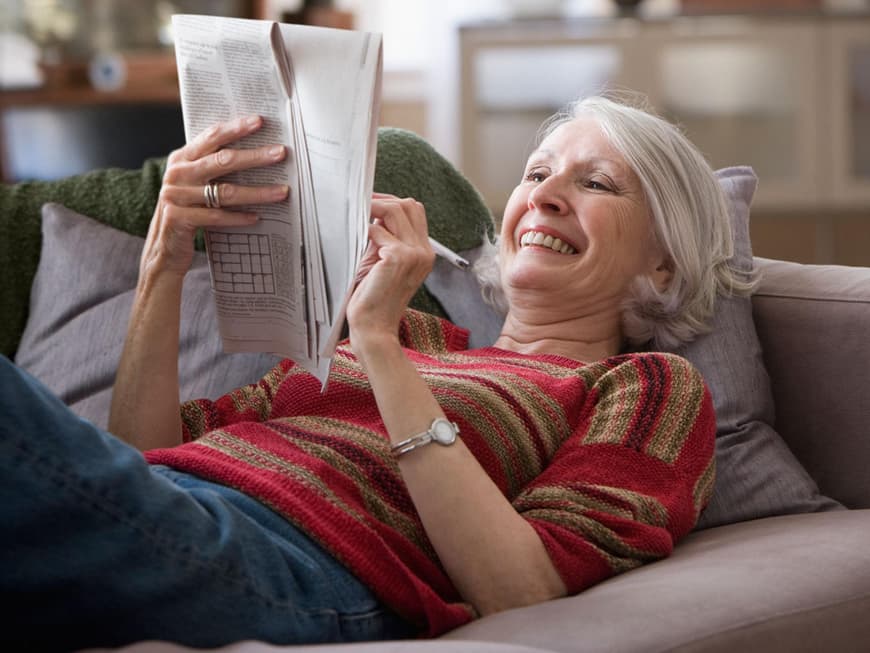 Seniorin liegt auf dem Sofa und löst Rätsel in Zeitung