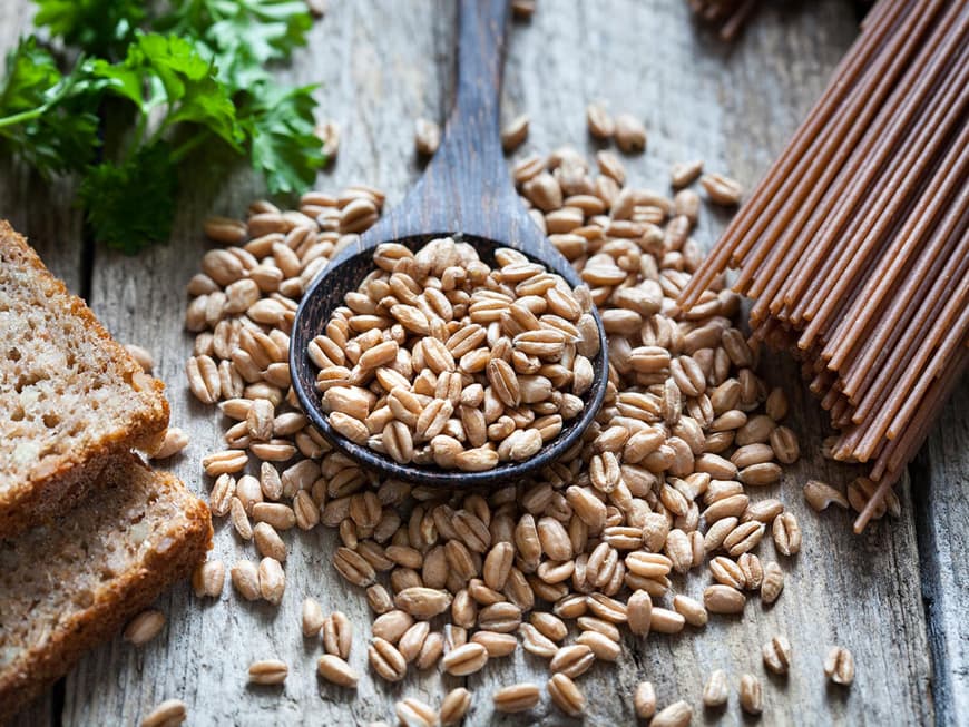 Vollkornbrot, Vollkorn-Nudeln und ein großer Löffel mit Körnern auf einem Holztisch