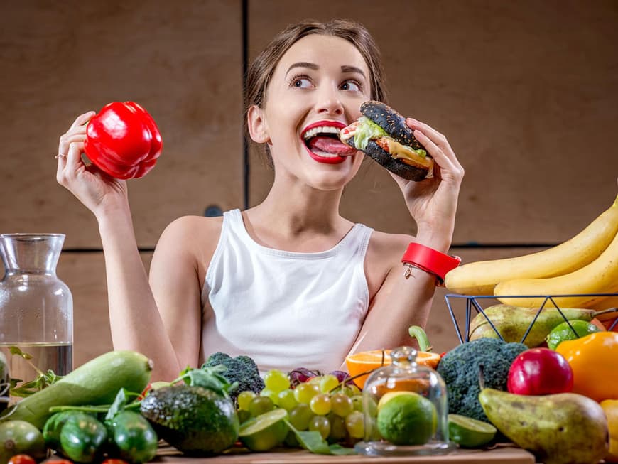 Frau sitzt an einem Tisch mit Gemüse, Obs und Getränken und hält eine Paprika und einen Burger in den Händen