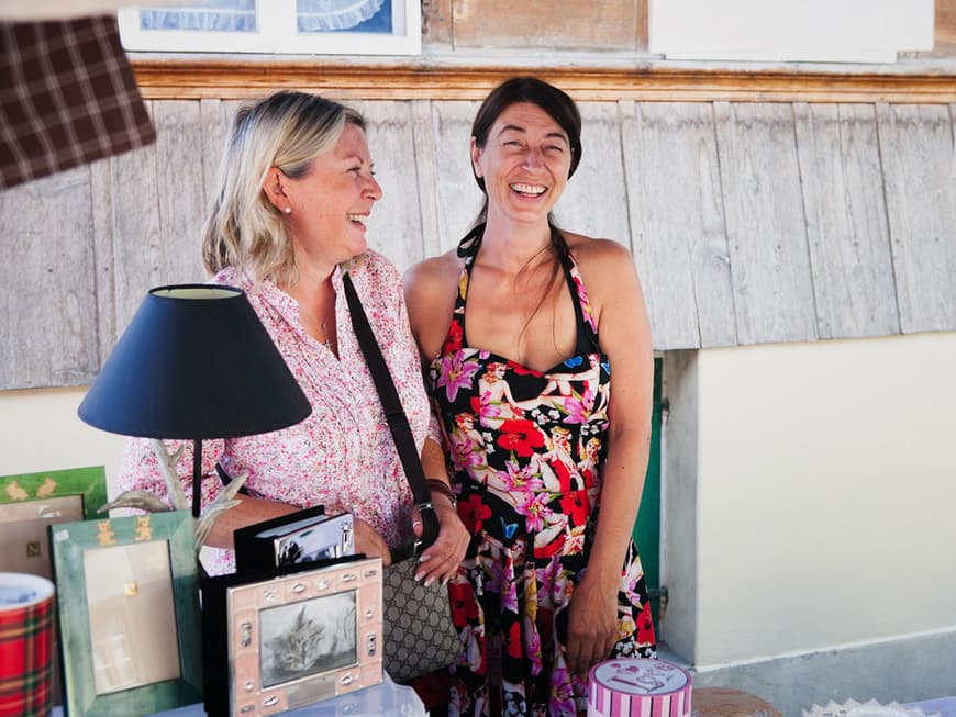 Zwei Frauen mittleren Alters an hinter einem Stand mit Bilderrahmen, Lampen etc.
