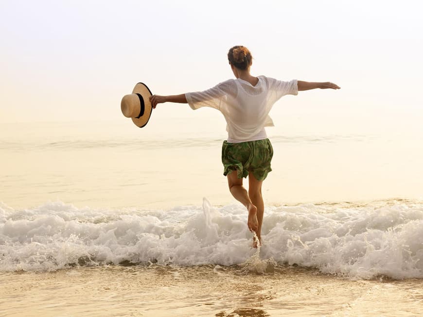 Frau mit Shorts und Bluse läuft mit offenen Armen auf das Meer zu, einen Hut in der Hand.