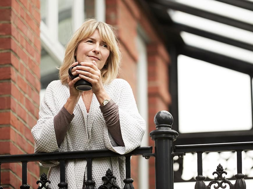 Blonde Frau mittleren Alters blickt mit Kaffeetasse in der Hand nachdenklich vom Balkon in die Ferne