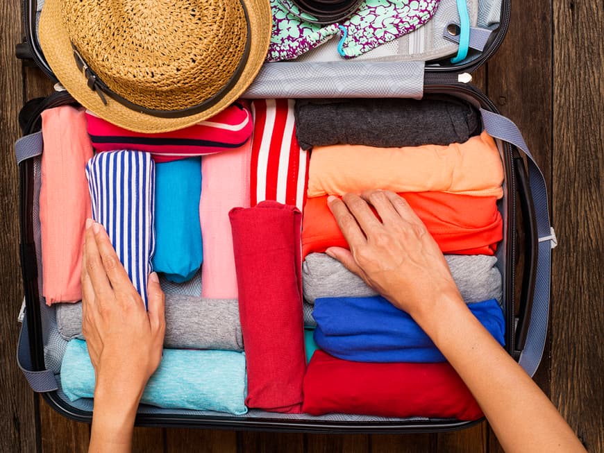 Koffer mit gerollten, bunten T-Shirts, einem Strohhut und einem Bikini