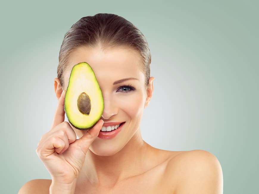Schöne Frau hält sich eine halbe Avocado vors Gesicht