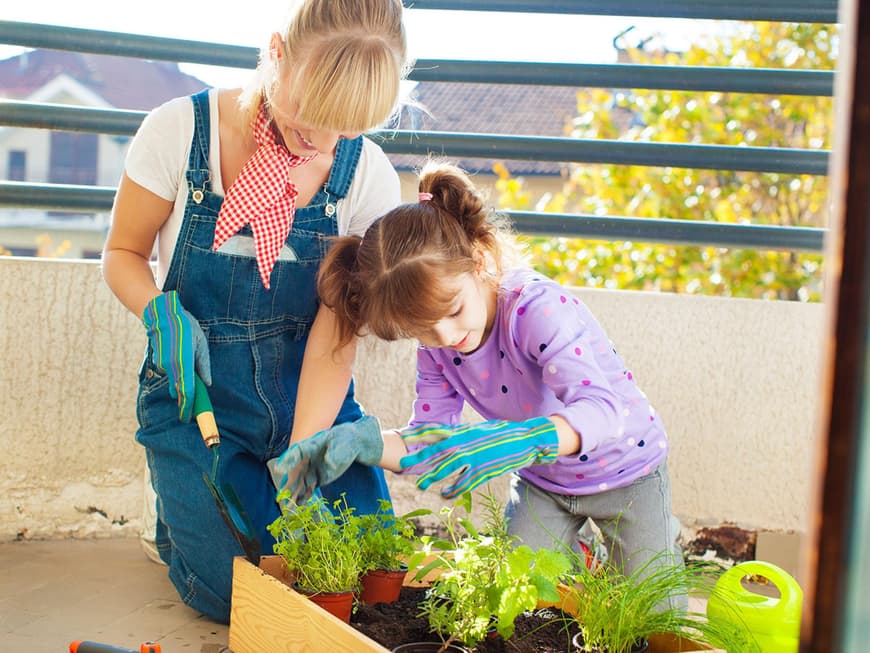 Mutter bepflanzt mit Tochter einen Blumenkasten mit Kräutern auf dem Balkon