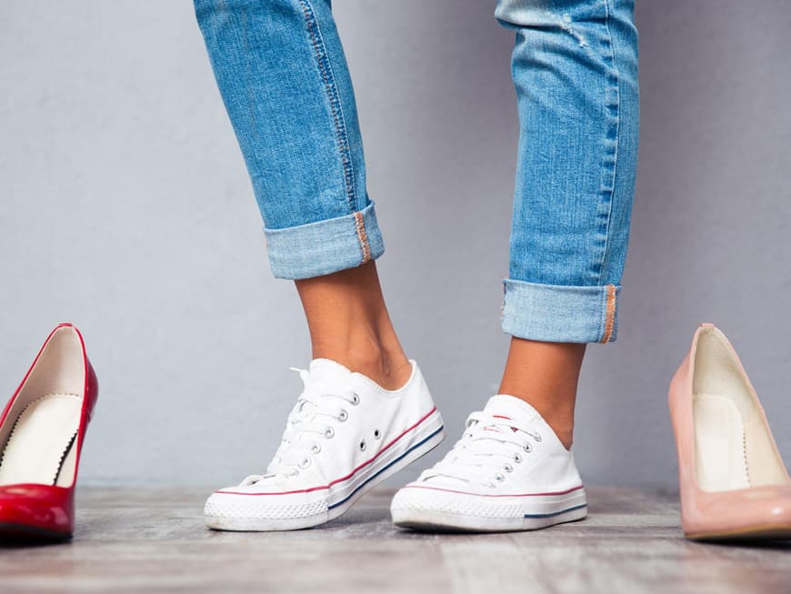 Frauenbeine in Jeans mit Sneakers neben verschiedenfarbigen Pumps