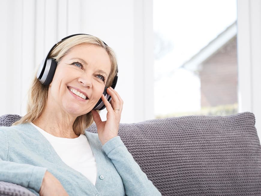 Ältere Frau sitzt auf dem Sofa und hört Musik über Kopfhörer